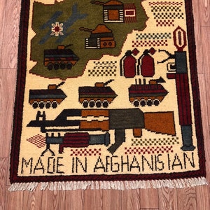 May include: A beige rug with a map of Afghanistan, military vehicles, and weapons woven into the design. The rug has a red border and fringe. The text "MADE IN AFGHANISTAN" and "2023" are woven into the rug.