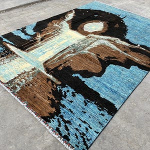 May include: A rectangular rug with an abstract design. The rug features a light blue background with brown, black, and cream-colored shapes. The edges are finished with a white fringe. The rug's design gives the impression of a watercolor painting.