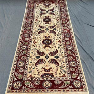 May include: A rectangular, cream-colored rug with a detailed, symmetrical pattern. The rug features a central design with floral and paisley motifs in burgundy, green, and blue, framed by a burgundy border. The rug has a fringe on both ends.