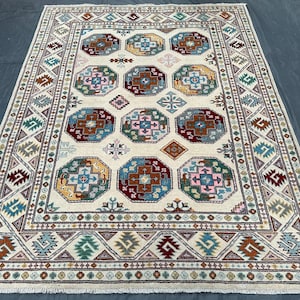 Könnte beinhalten: Rechteckiger beigefarbener Teppich mit geometrischem Muster. Der Teppich hat ein zentrales Design aus Achtecken in Blau-, Rot-, Rosa- und Gelbtönen, umgeben von einem gemusterten Rand in komplementären Farben. Geeignet für die Inneneinrichtung.