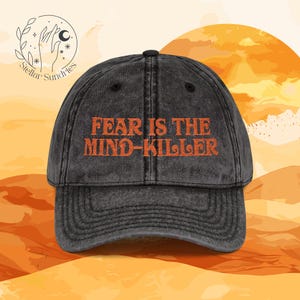May include: A distressed black baseball cap with the orange text "FEAR IS THE MIND-KILLER." The cap has a curved brim and a button on top. The background features a desert landscape with a sun and the Stellar Sundries logo.