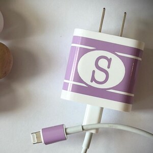 May include: A purple phone charger with a white oval containing the letter "S" on the front. The charger is plugged into a white cable with a purple lightning connector.