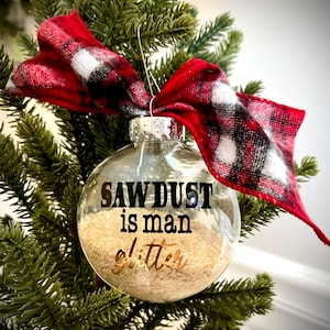 May include: Clear glass Christmas ornament filled with sawdust and the text "SAW DUST is man glitter" in black and gold lettering. The ornament is adorned with a red and black plaid ribbon and is hanging on a green Christmas tree.