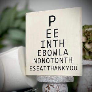 May include: White wooden sign with black text that reads "Pee in the bowl and not on the seat thank you". The sign is sitting on a white pedestal.