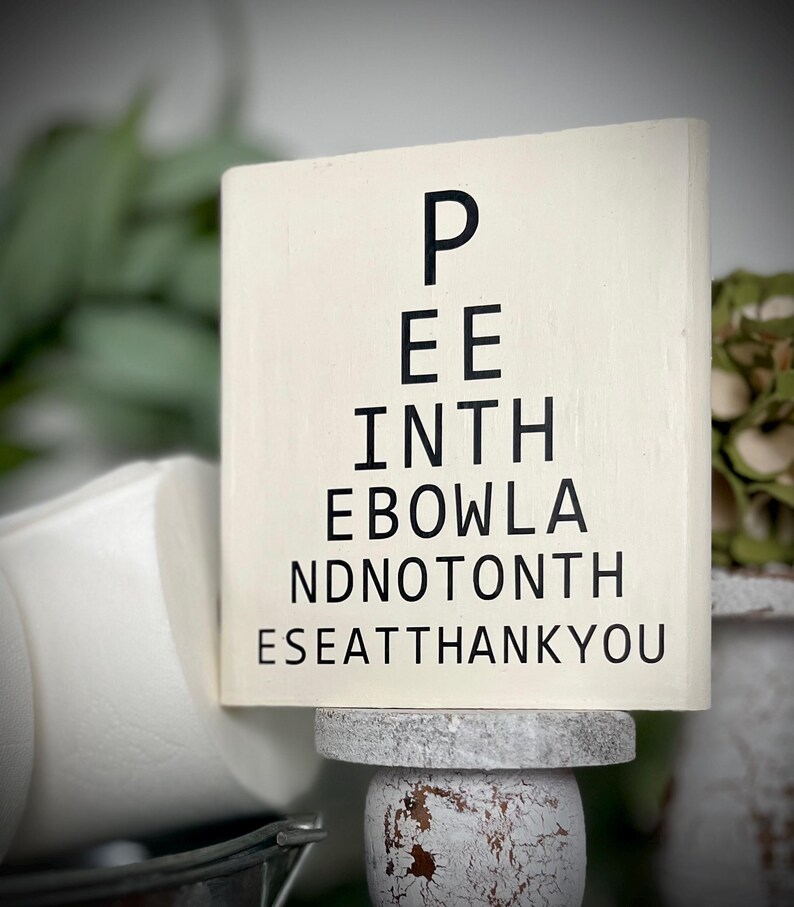 May include: White wooden sign with black text that reads "Pee in the bowl and not on the seat thank you". The sign is sitting on a white pedestal.