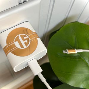 May include: A white phone charger with a gold monogram sticker on the side. The charger is plugged into an outlet and a white charging cable is connected to the charger.