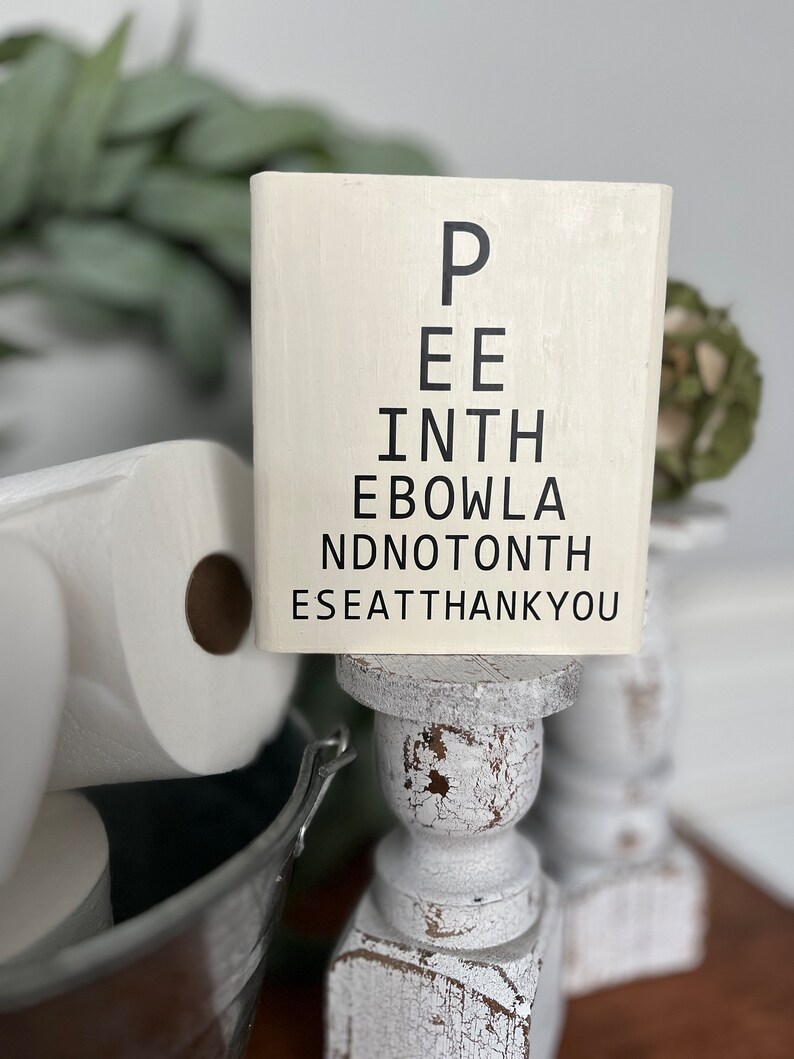 May include: White wooden sign with black text that reads "Pee in the bowl and not on the seat thank you". The sign is on a white wooden pedestal.