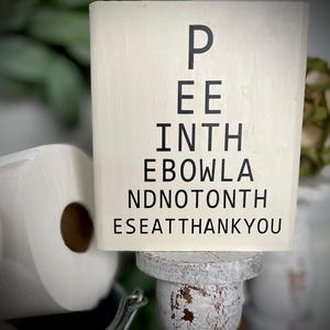 May include: White wooden sign with black text that reads "Pee in the bowl and not on the seat thank you".
