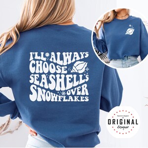 May include: A blue sweatshirt with white text that reads "I'll Always Choose Seashells Over Snowflakes." A seashell and snowflake graphic is included in the design. A smaller seashell graphic is on the upper right chest.