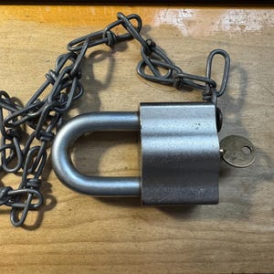 May include: A silver-toned metal padlock with a thick, rounded shackle and a matching metal chain attached to the top. A small, circular brass-colored key is inserted into the side of the lock body, which features a smooth, brushed finish.