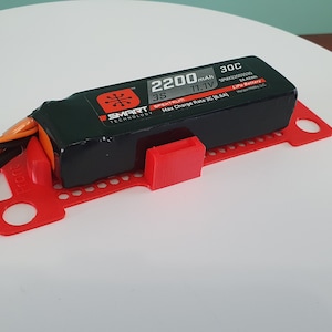 May include: A red 3D printed battery holder with a black 2200 mAh LiPo battery. The battery has the text "SMART" and "TECHNOLOGIES" on it. The battery is connected to wires.