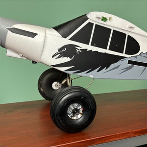 Puede incluir: Un avión modelo blanco y gris con detalles negros y un gráfico de cuervo. El avión tiene una escena de montaña en blanco y negro en el lateral y el texto "N3". El avión tiene tres ruedas, dos de las cuales son visibles en la imagen.