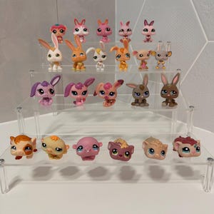 May include: A collection of colorful, small animal figurines, including rabbits and other creatures, displayed on a clear, tiered shelf. The figurines feature various colors like pink, orange, brown, and white, with detailed facial features and designs.