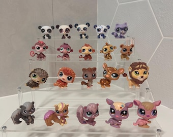 Puede incluir: Colección de pequeñas figuras de animales coloridos, incluyendo pandas, monos y ciervos, exhibidas en una estantería transparente de varios niveles. Las figuras tienen ojos grandes y detalles, con una variedad de colores y patrones.