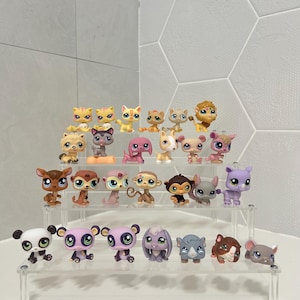 May include: A collection of twenty-three small plastic animal figurines with oversized eyes, arranged on a three-tiered clear acrylic display stand. The figures include various species such as cats, dogs, monkeys, a panda, and a lion in diverse colors.