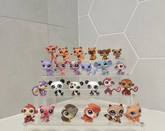 May include: A collection of twenty-three small, stylized animal figurines arranged on a clear, three-tiered acrylic display stand. The figures include various species such as monkeys, pandas, a giraffe, a raccoon, and a hedgehog, featuring large eyes.