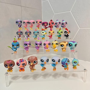 May include: A collection of twenty-eight small, stylized bird figurines arranged on a clear, four-tiered acrylic display stand. The figures feature diverse color palettes, including pink, blue, yellow, brown, and purple, with large, expressive eyes.