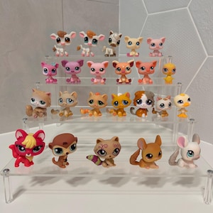 Puede incluir: Una colección de pequeñas figuras de animales de plástico de colores, incluyendo gatos, perros y otras criaturas, se exhibe en un expositor transparente de varios niveles. Las figuras presentan varios colores y diseños, con ojos grandes y poses juguetonas.