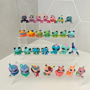 May include: A collection of small, colourful plastic animal figurines, including frogs and other creatures, displayed on clear acrylic shelves. The figurines feature various colours and designs, with some wearing accessories like bows and hats. The arrangement is on a white surface.