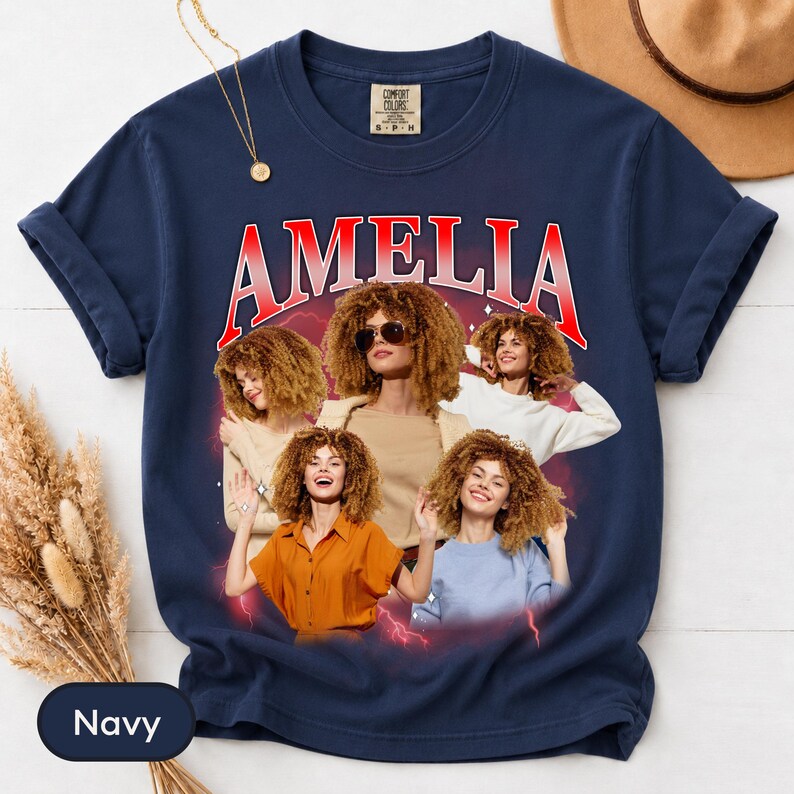 May include: A navy blue t-shirt featuring the name AMELIA in red and white arched lettering. The graphic includes four photographic portraits of a person with curly hair in various poses and outfits, set against a dark red background with lightning.