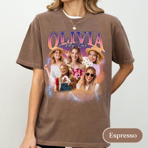 May include: An espresso-coloured t-shirt featuring a graphic print with the text OLIVIA My Love. The design includes a collage of four individuals in various poses, accented by a purple and orange gradient background and blue lightning bolt graphics.
