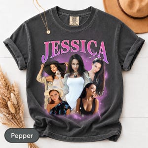 May include: A charcoal grey short-sleeve t-shirt featuring the name JESSICA in large pink letters. The graphic includes a collage of four different images of the same person in various outfits, surrounded by a purple lightning effect.