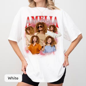 May include: A white short-sleeve t-shirt featuring a graphic print with the name AMELIA in red and silver lettering at the top. The design includes a collage of four images of a person with curly hair wearing different outfits and sunglasses.