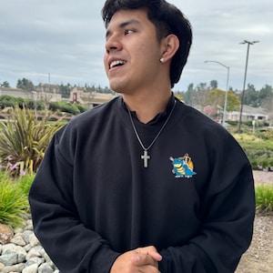 May include: Black sweatshirt with a small cartoon graphic on the front. The sweatshirt has a crew neck and long sleeves. A silver cross pendant hangs from a chain around the neck. The person is wearing a black ring.