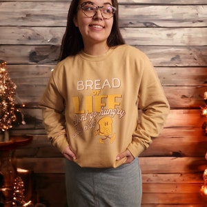 May include: A mustard yellow sweatshirt with the text "Bread of Life" and a cartoon bread character. The text below the character reads "Don't go hungry John 6:35".