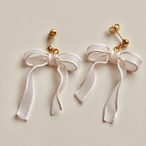 May include: A pair of white ribbon bow earrings with gold-toned metal findings.