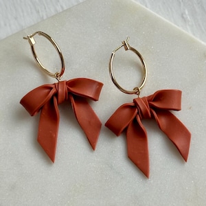 May include: A pair of terracotta-colored bow earrings with gold-tone hoop closures. The bows are a matte finish and have a classic bow shape. The earrings are displayed on a white marble surface.