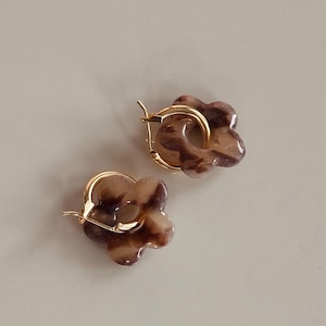 Könnte beinhalten: Ein Paar blütenförmige Ohrringe. Jeder Ohrring hat einen goldfarbenen Bügel und ein marmoriertes, blütenblattartiges Design in Braun- und Cremetönen. Die Ohrringe werden auf einer neutralen, hellfarbigen Oberfläche präsentiert.