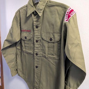 May include: Olive green button-up shirt with two chest pockets and a collar. The shirt has the text "BOY SCOUTS OF AMERICA" embroidered in red above the left pocket and a "SULLIVAN COU" patch on the right shoulder.