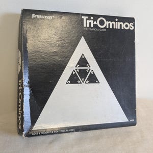May include: A black and silver Tri-Ominos game box. The box features a large white triangle with smaller triangles inside, each with numbers. The game's name is in white text. The box is designed for ages 8 to adult and for 2 to 6 players.