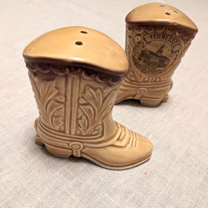 May include: A pair of ceramic cowboy boot salt and pepper shakers. The shakers are brown and white with a detailed design. One shaker has the words "Tennessee" and "Capitol Building" on it.