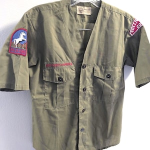 May include: Olive green Boy Scouts of America short-sleeve shirt with two chest pockets and button closures. Features patches on the sleeves with a horse and the words "Big Horn" and "Sullivan Co."