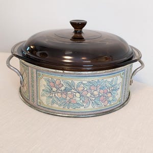 May include: A vintage casserole dish with a clear, brown glass lid and a metal base. The base features a decorative floral design with pink, yellow, and blue fruits and leaves. The dish has metal handles on either side.