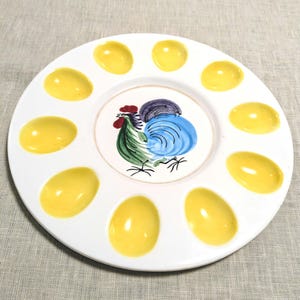 May include: White ceramic deviled egg serving platter with a hand-painted rooster design in the center. The platter has 12 yellow egg-shaped indentations around the perimeter. A decorative and functional serving piece.
