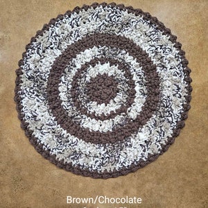 May include: A brown and white crocheted round rug with an ombre effect. The rug is 15 inches in diameter and has a spiral pattern. Brown/Chocolate Ombre 15" is written below the rug.