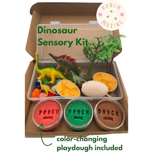 May include: A Dinosaur Sensory Kit in an open cardboard box. The kit includes dinosaur figurines, a small plastic tree, and three containers of color-changing playdough. The text "Doughmazing" is on a round sticker. The text "color-changing playdough included" is also visible.