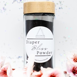 Op de afbeelding: Een glazen pot met een houten deksel gevuld met zwart poeder. De pot heeft een wit etiket met de tekst "Diaper Bliss Powder Activated Coconut Charcoal" en een kleiner etiket met een berglogo waarop "soul blends" staat.