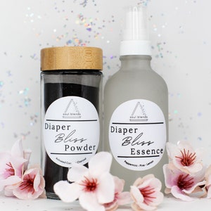 Könnte beinhalten: Zwei Glasflaschen mit schwarzen und weißen Etiketten. Die Flasche links ist ein schwarzes Pulver mit der Aufschrift "Diaper Bliss Powder - Activated Charcoal - Charcoal". Die Flasche rechts ist eine transparente Sprühflasche mit der Aufschrift "Diaper Bliss Essence - Peppermint - Pine - Lemon".