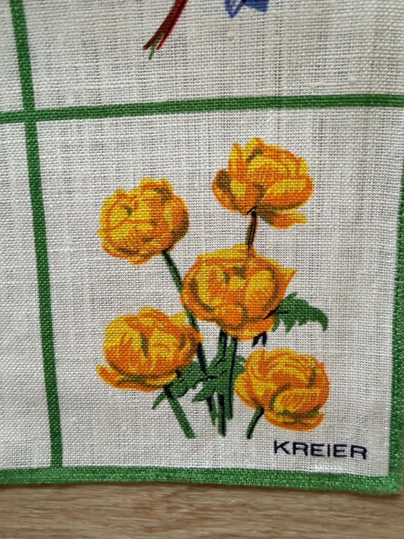 May include: A white linen handkerchief with a green border and grid pattern. The handkerchief features a cluster of orange and yellow flowers with green stems and leaves. The word "KREIER" is printed in black.