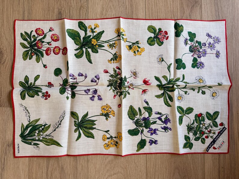 May include: A white linen handkerchief with a red border, adorned with floral illustrations in red, yellow, purple, and white. The design features various flowers and green leaves, creating a botanical theme.