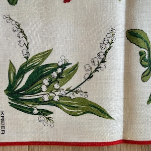 May include: A linen textile with a red border features a floral design of green leaves and white bell-shaped flowers. The text "KREIER" is printed in black. The background is a light beige color.