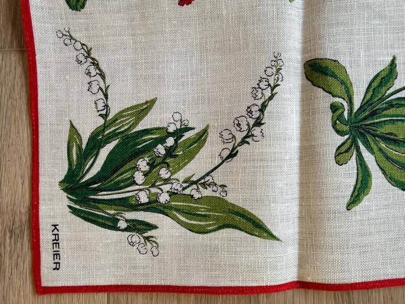 May include: A linen textile with a red border features a floral design of green leaves and white bell-shaped flowers. The text "KREIER" is printed in black. The background is a light beige color.