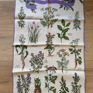 May include: A beige linen kitchen towel featuring illustrations of various herbs with their names in German. The towel has a purple border and a purple banner that reads "Herbs - Gew&uuml;rze". The towel includes illustrations of chives, thyme, and dill.