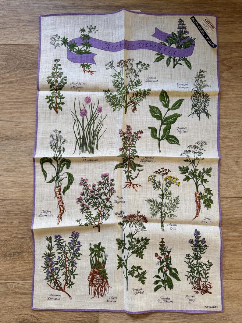May include: A beige linen kitchen towel featuring illustrations of various herbs with their names in German. The towel has a purple border and a purple banner that reads "Herbs - Gew&uuml;rze". The towel includes illustrations of chives, thyme, and dill.