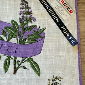 May include: Linen kitchen towel with a purple border and a botanical illustration. The design features green leaves, purple flowers, and a purple ribbon. The towel has a tag that reads "KREIER" and "REINLEINEN - PUR FIL".
