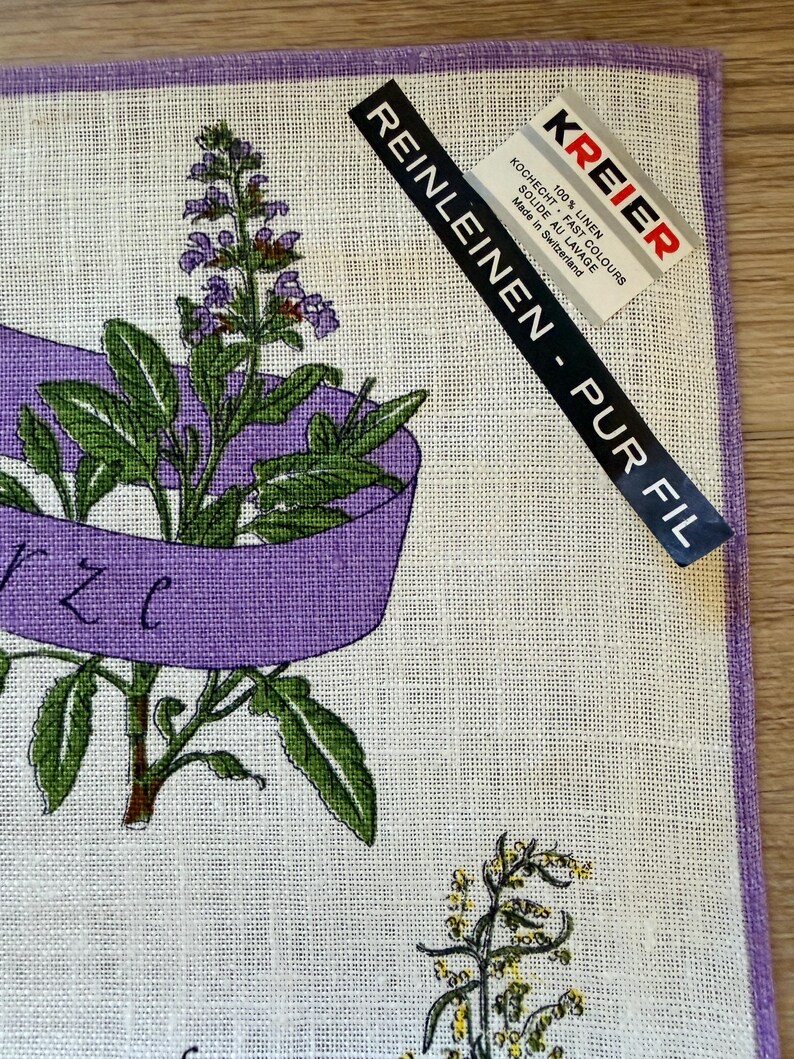 May include: Linen kitchen towel with a purple border and a botanical illustration. The design features green leaves, purple flowers, and a purple ribbon. The towel has a tag that reads "KREIER" and "REINLEINEN - PUR FIL".